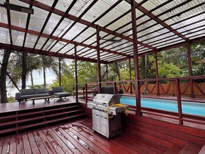 A covered outdoor area with a pool, wooden deck, grill, and seating.