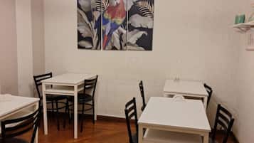 Uma sala de jantar com mesas brancas e cadeiras pretas, parede com pintura de um papagaio, e uma estante com objetos.