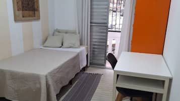 Quarto pequeno com uma cama de solteiro, escrivaninha e cadeira, janela com persianas e vista para o exterior.