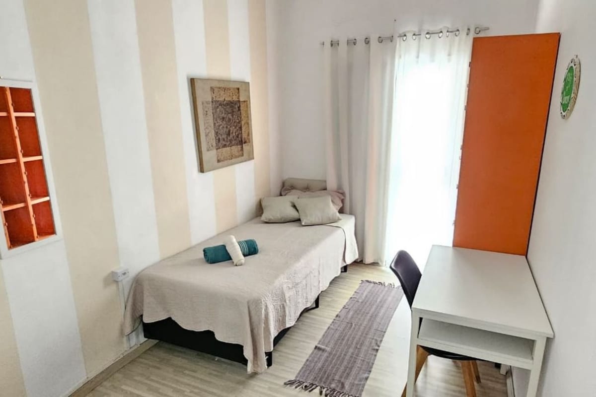 A bedroom with a bed, a desk, a chair, a window with curtains, and a wall-mounted artwork.