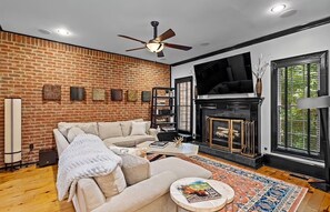 A living room with a brick wall, a fireplace, a ceiling fan, and a large window. - Furnished Executive Townhome - Dedicated WFH Office - Ideal for 6+ Month Stays (Atlanta)