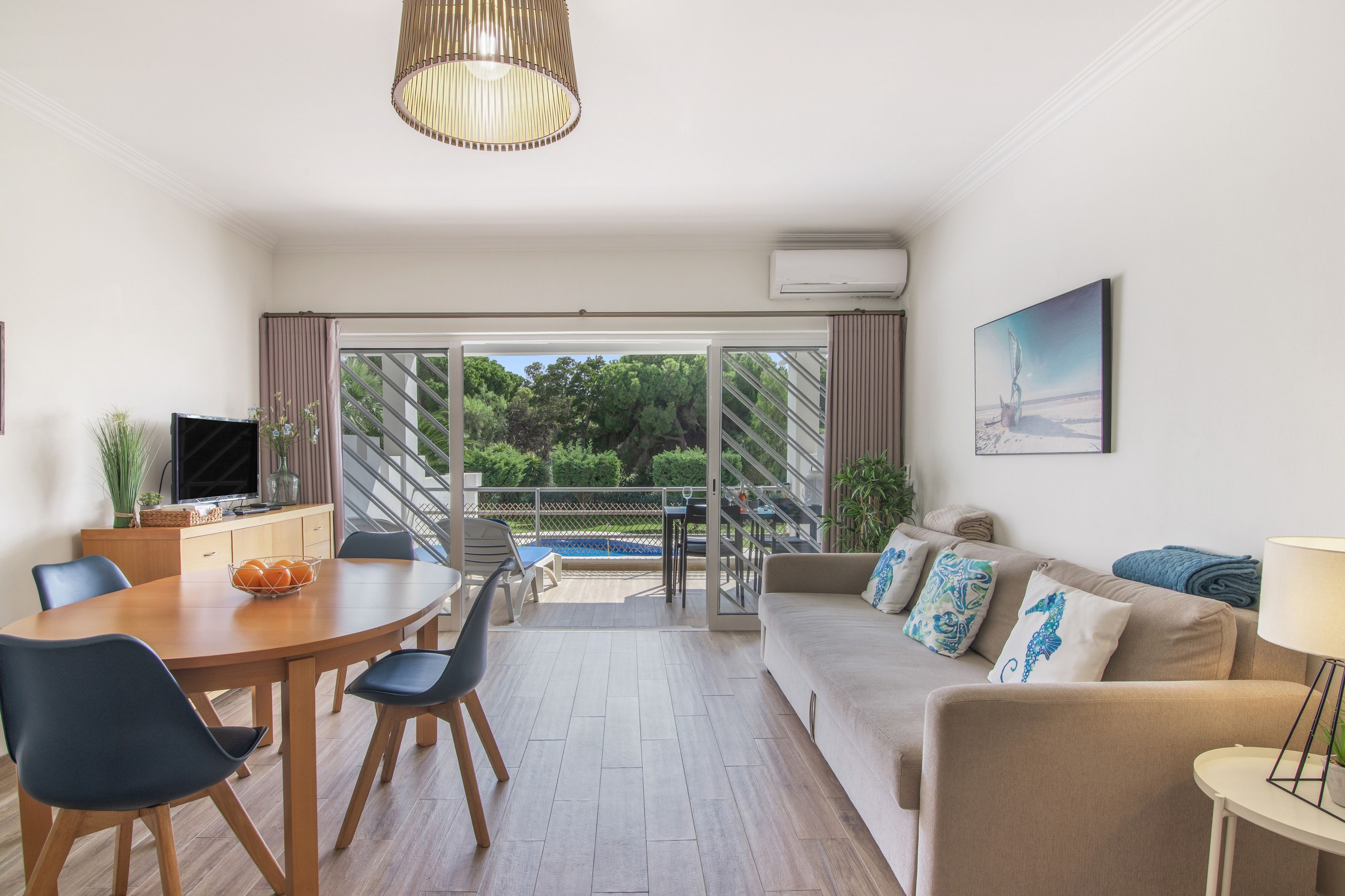 Una sala de estar moderna con comedor, un sofá con almohadones decorativos, un televisor y vistas a una piscina y follaje exterior.
