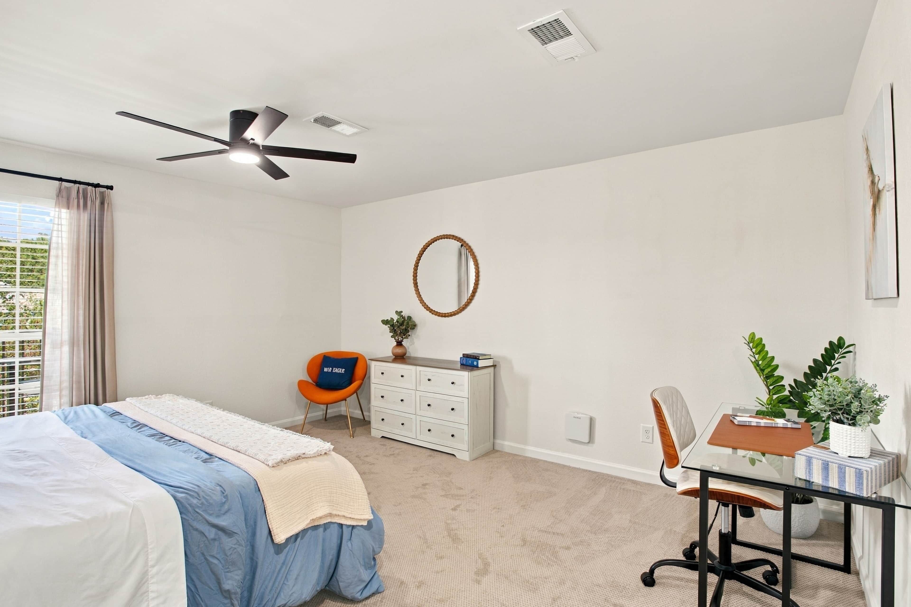 Un dormitorio con cama, escritorio, silla, ventilador de techo y ventana con cortinas.