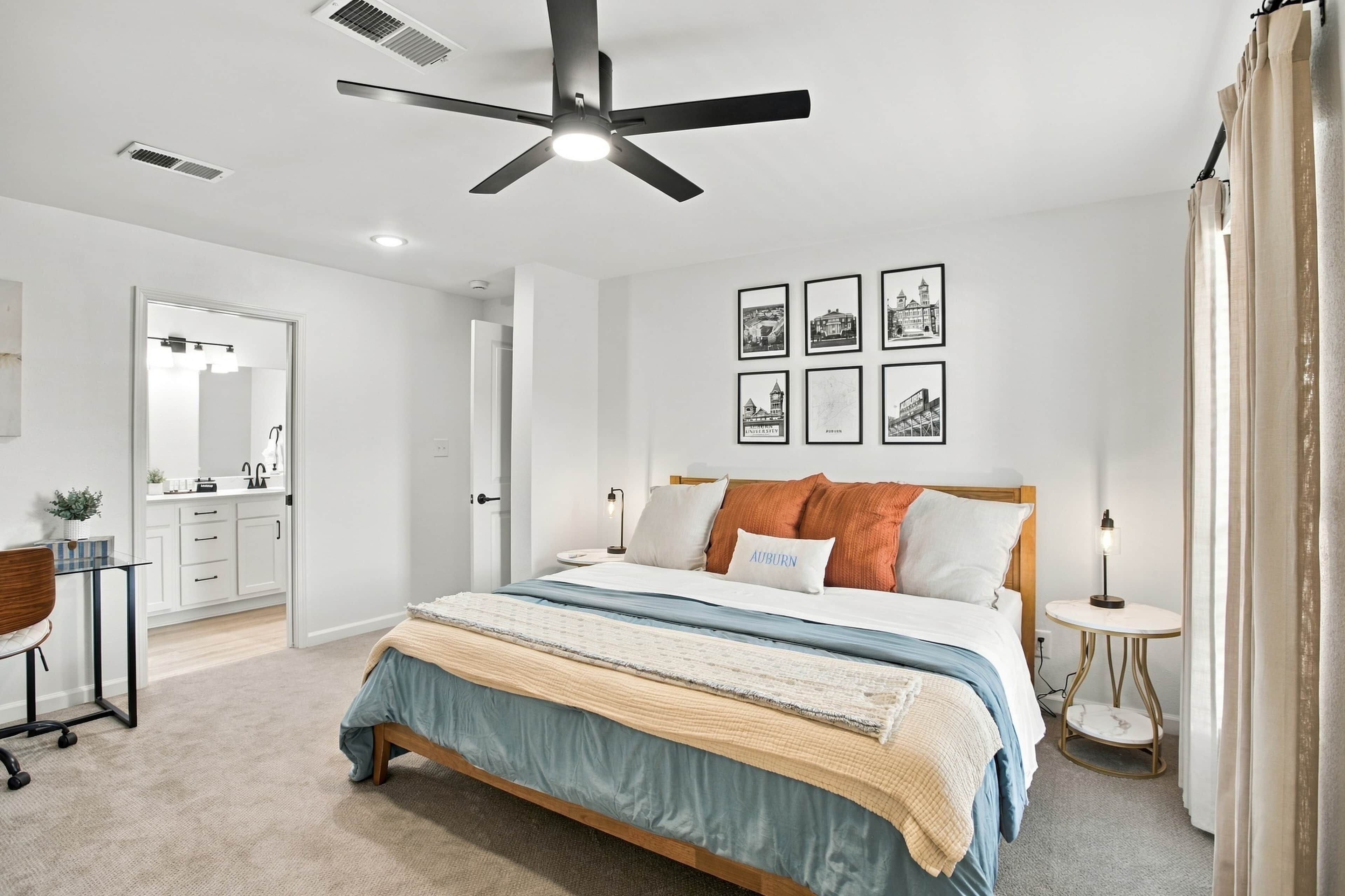 A bedroom with a bed, a ceiling fan, a nightstand, a lamp, and framed pictures on the wall.