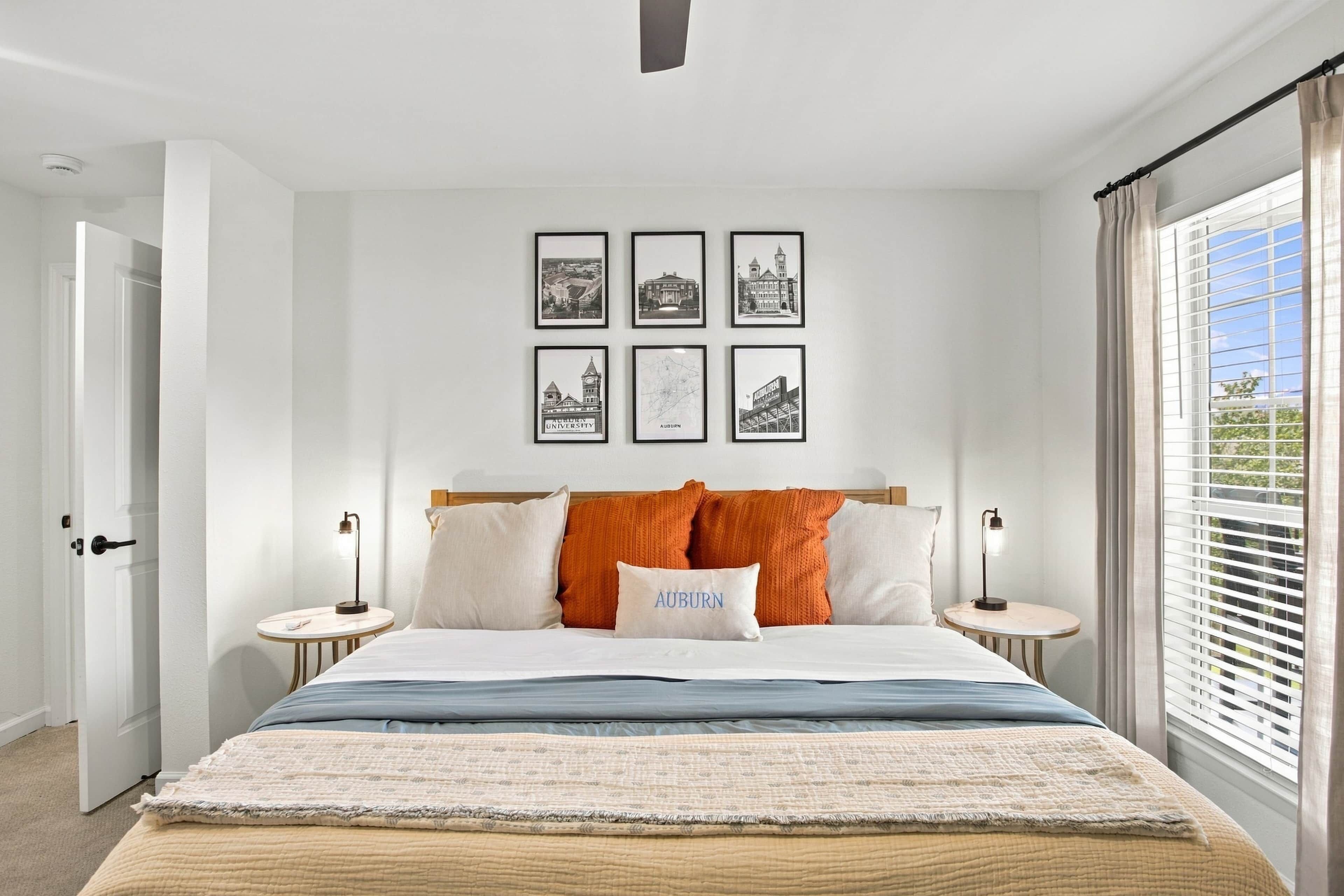 A bedroom with a bed, a nightstand, a wall-mounted lamp, a window with blinds, and framed pictures on the wall.