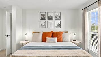 A bedroom with a bed, a nightstand, a wall-mounted lamp, a window with blinds, and framed pictures on the wall.