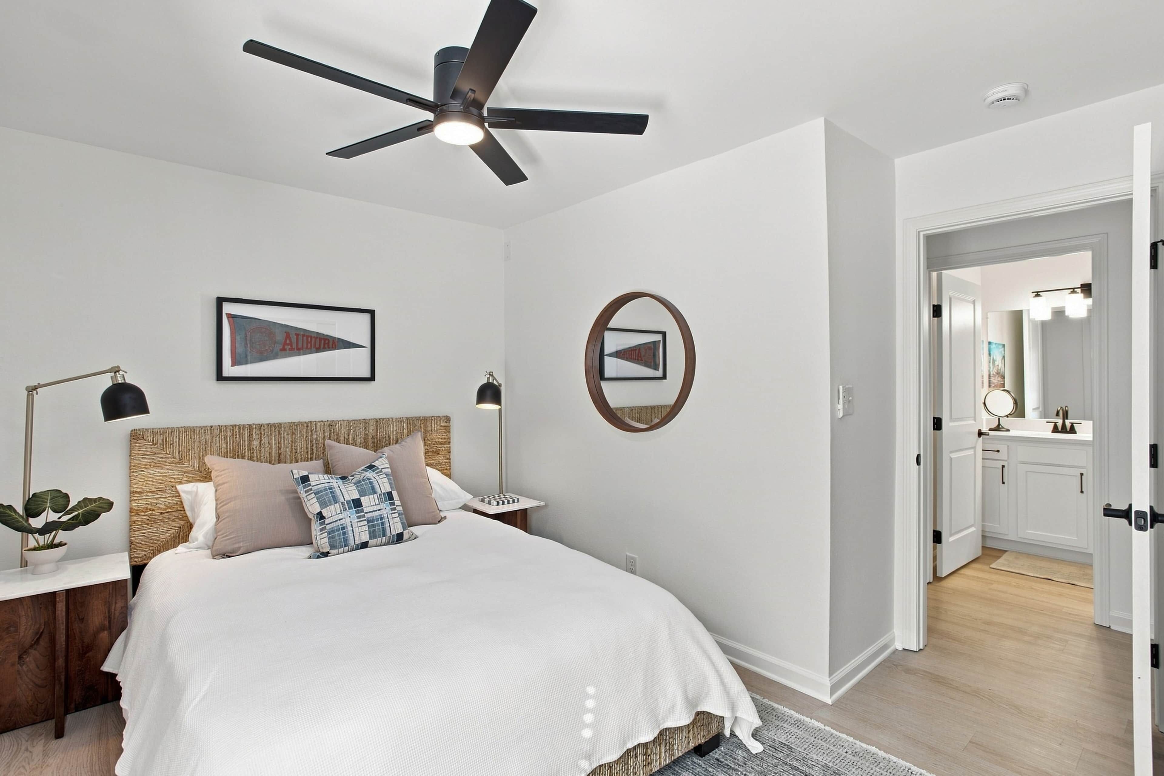 A bedroom with a bed, a ceiling fan, a nightstand, a lamp, a framed picture, a mirror, and a door leading to a bathroom.