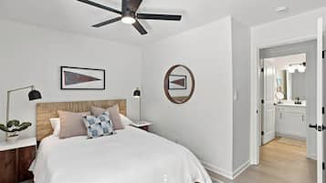 A bedroom with a bed, a ceiling fan, a nightstand, a lamp, a framed picture, a mirror, and a door leading to a bathroom.