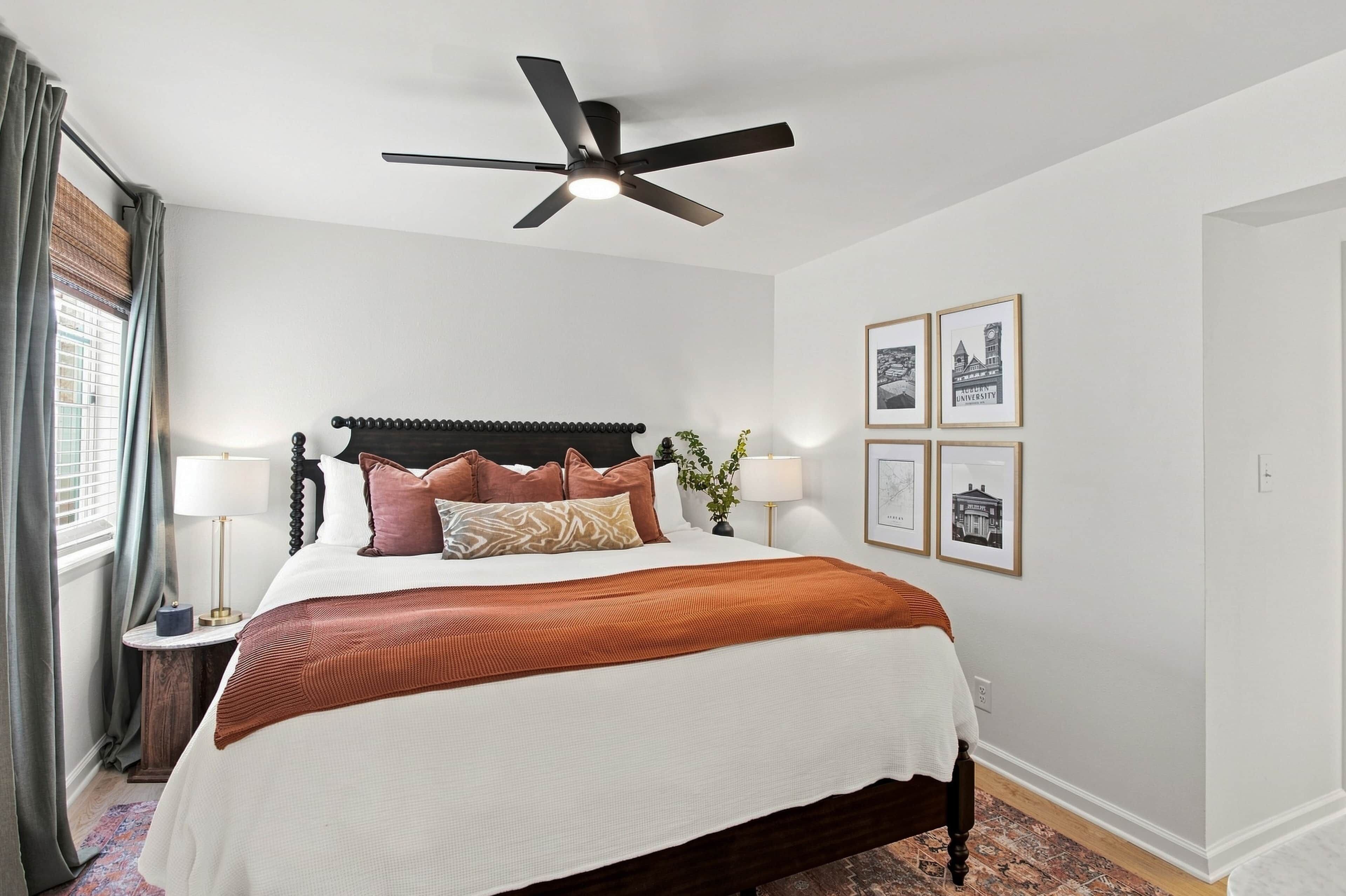 A hotel room with a bed, a ceiling fan, a nightstand with a lamp, and framed pictures on the wall.