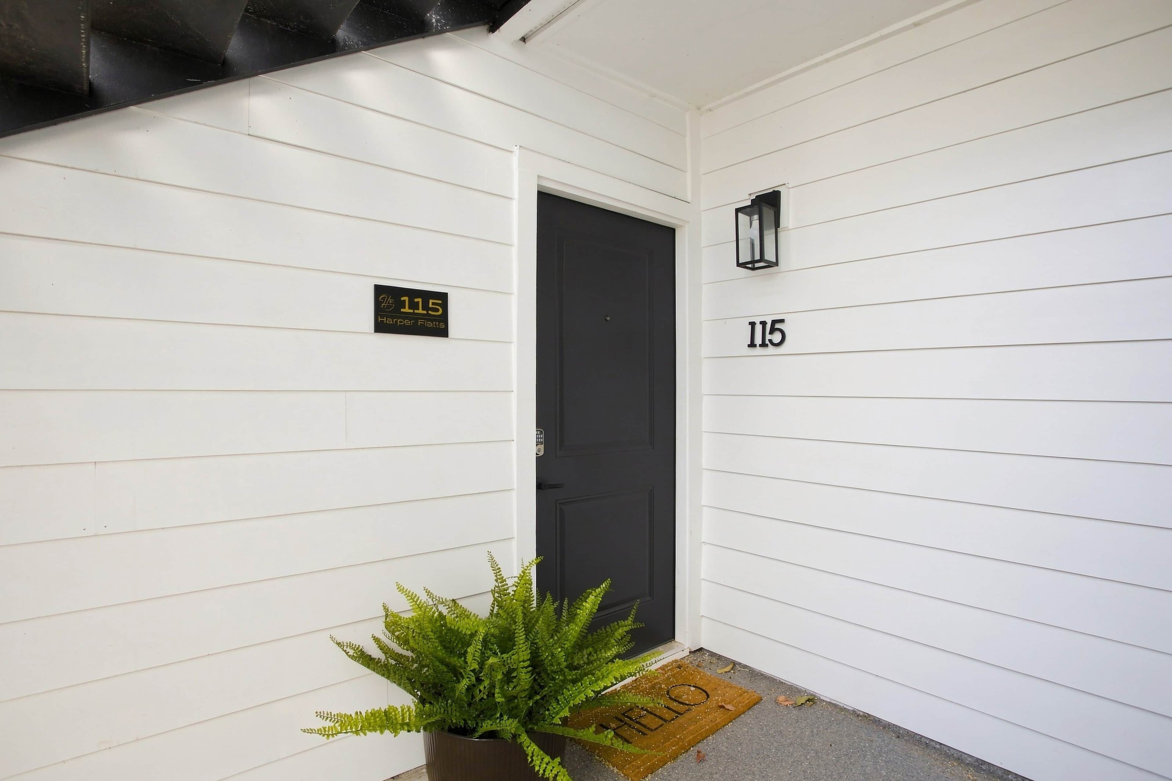 A white exterior wall with a black door, a number plaque, and a small wall-mounted light fixture.