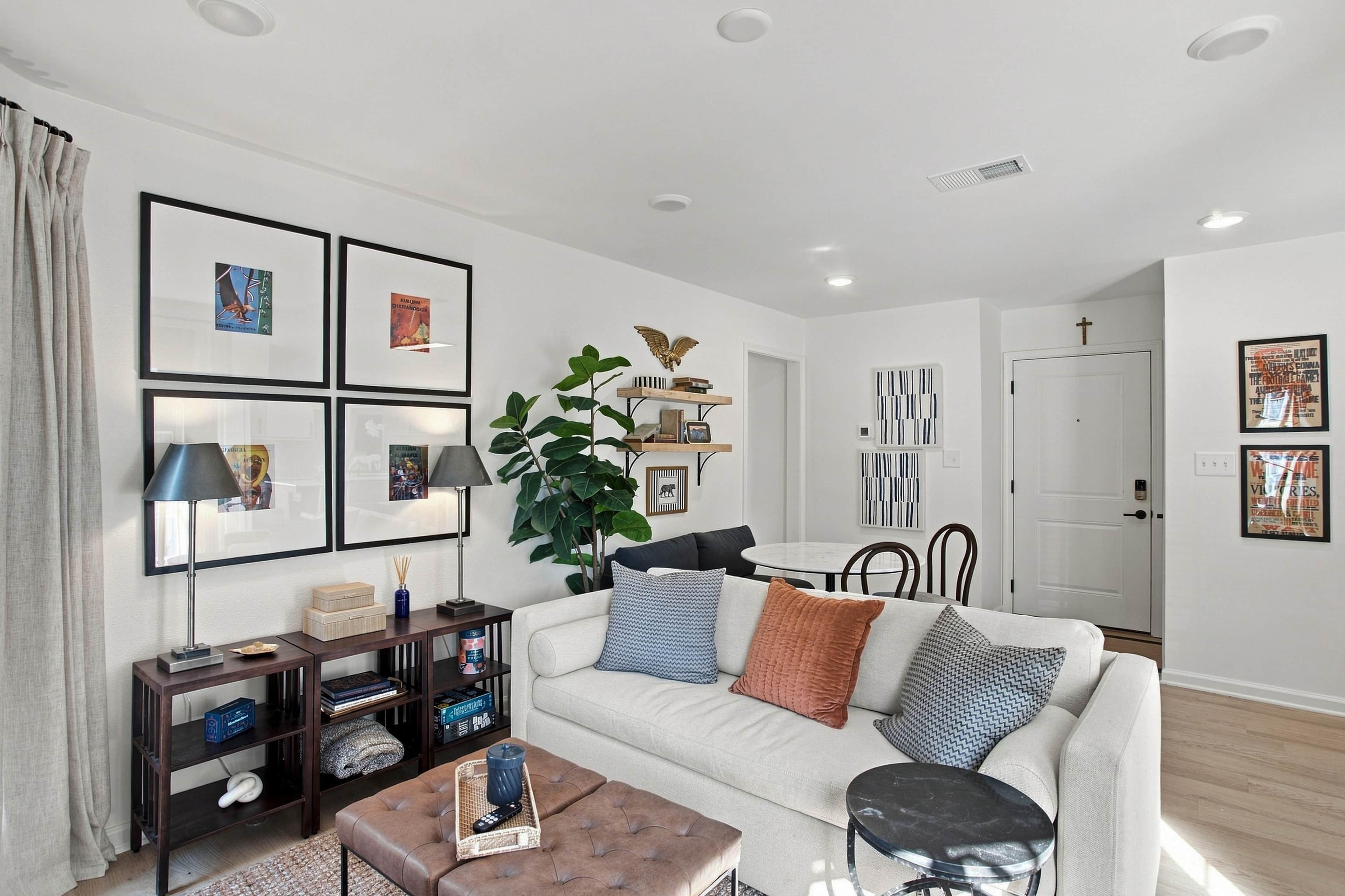 A modern living room with a white sofa, a coffee table, and a bookshelf. There are framed pictures on the wall and a plant on a side table.
