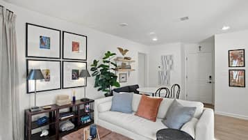 A modern living room with a white sofa, a coffee table, and a bookshelf. There are framed pictures on the wall and a plant on a side table.