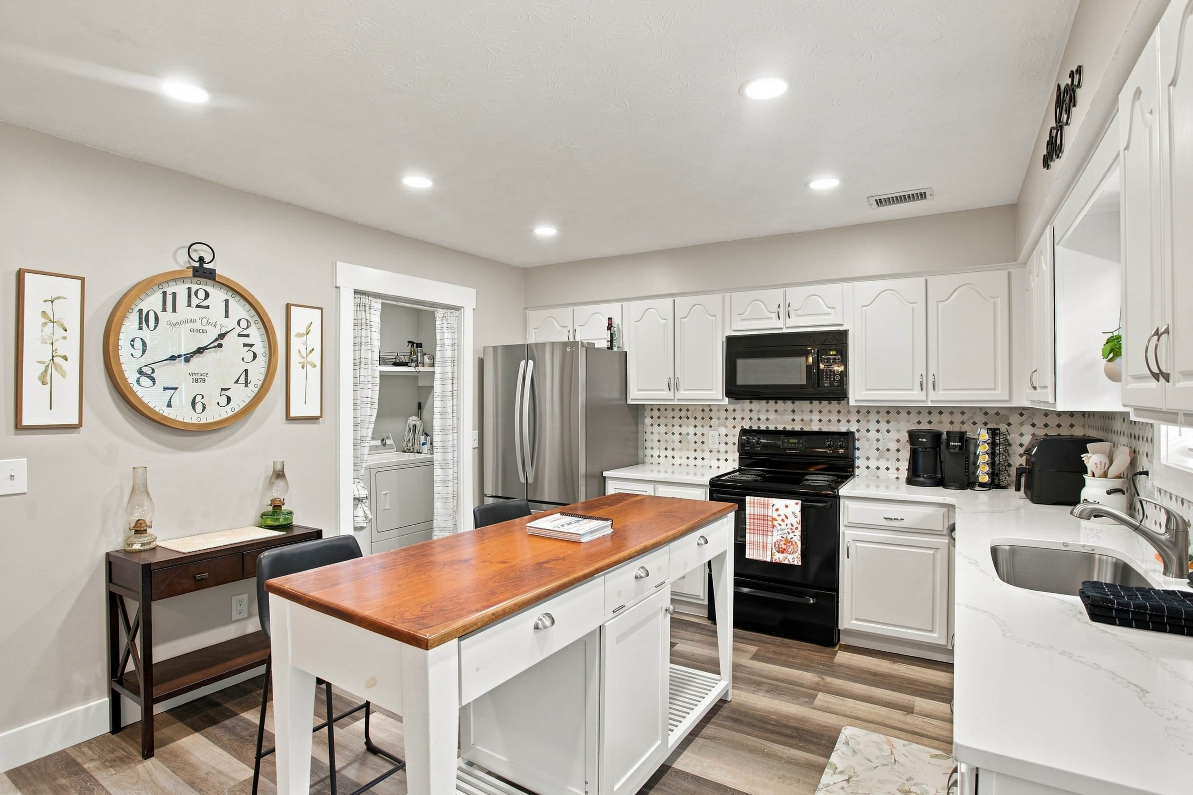 Una cocina moderna con armarios blancos, una isla de madera, electrodomésticos de acero inoxidable y un reloj de pared de estilo vintage.