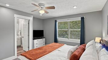 A bedroom with a bed, a television, a window with blinds, a ceiling fan, and a door leading to another room.