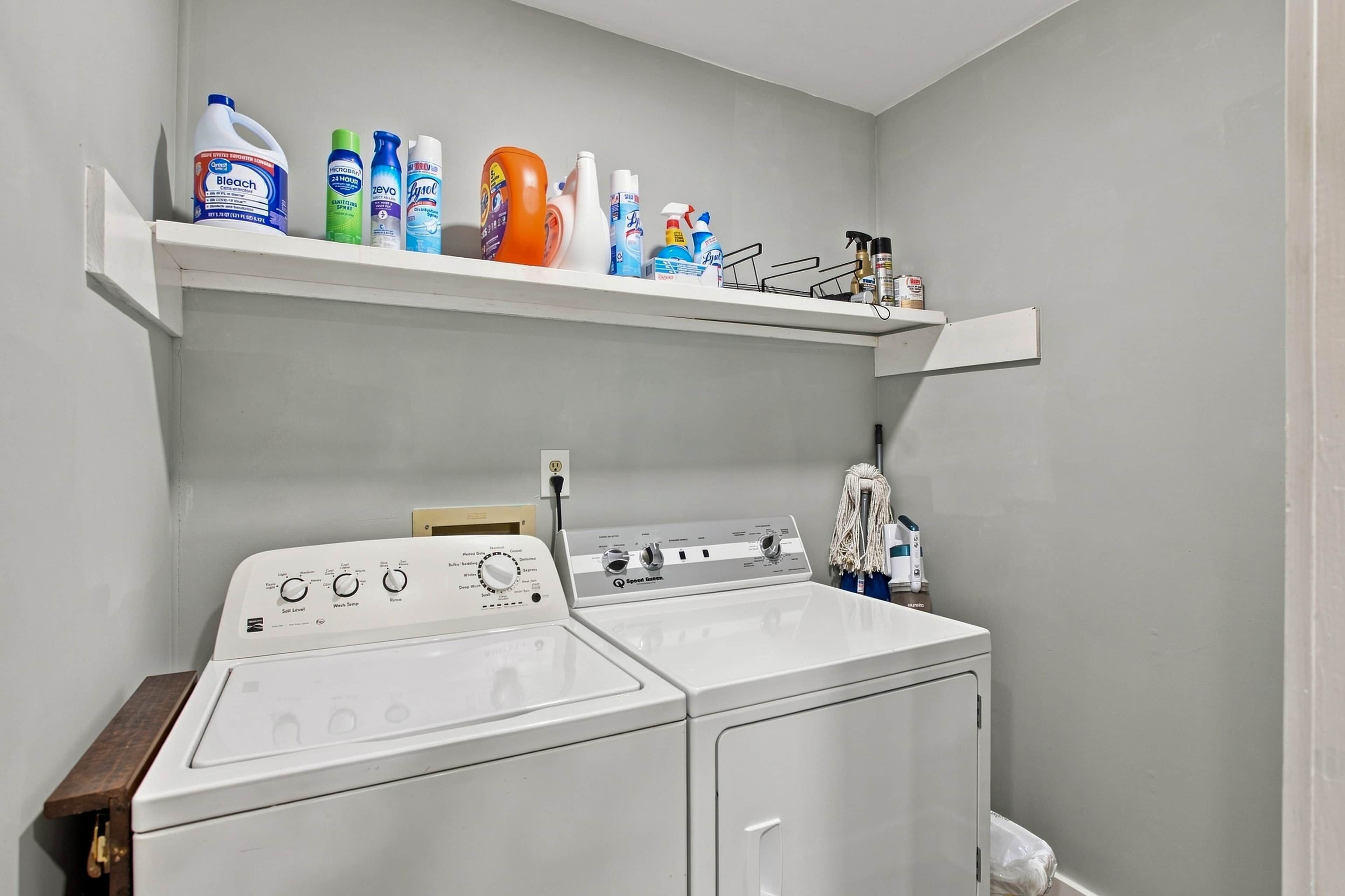 A laundry area with a front-loading washing machine, a shelf above it holding various cleaning products, and a wall-mounted power outlet.