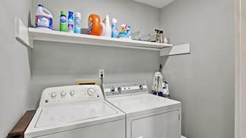 A laundry area with a front-loading washing machine, a shelf above it holding various cleaning products, and a wall-mounted power outlet.