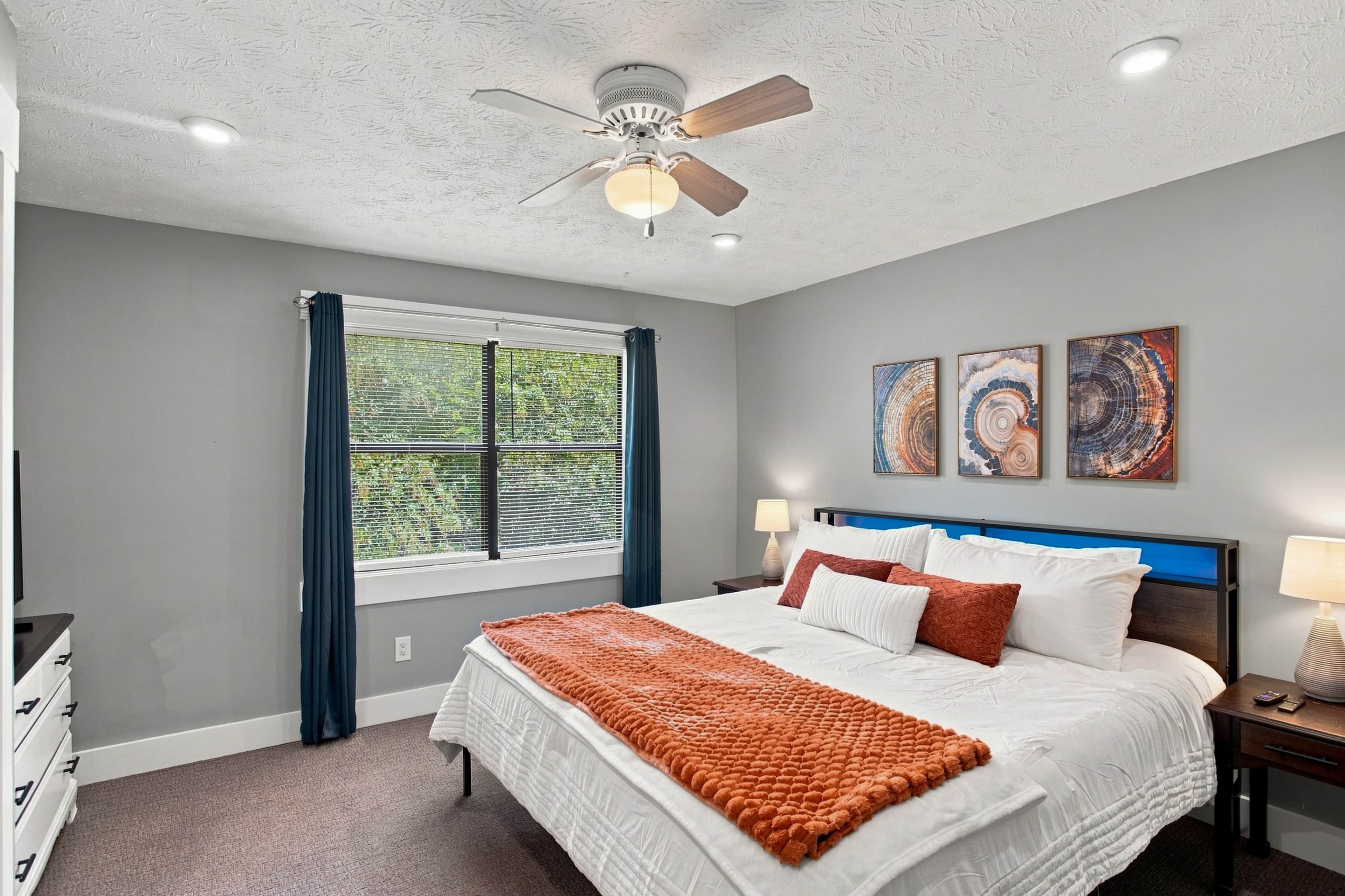 A bedroom with a bed, a ceiling fan, a window with curtains, and wall art.