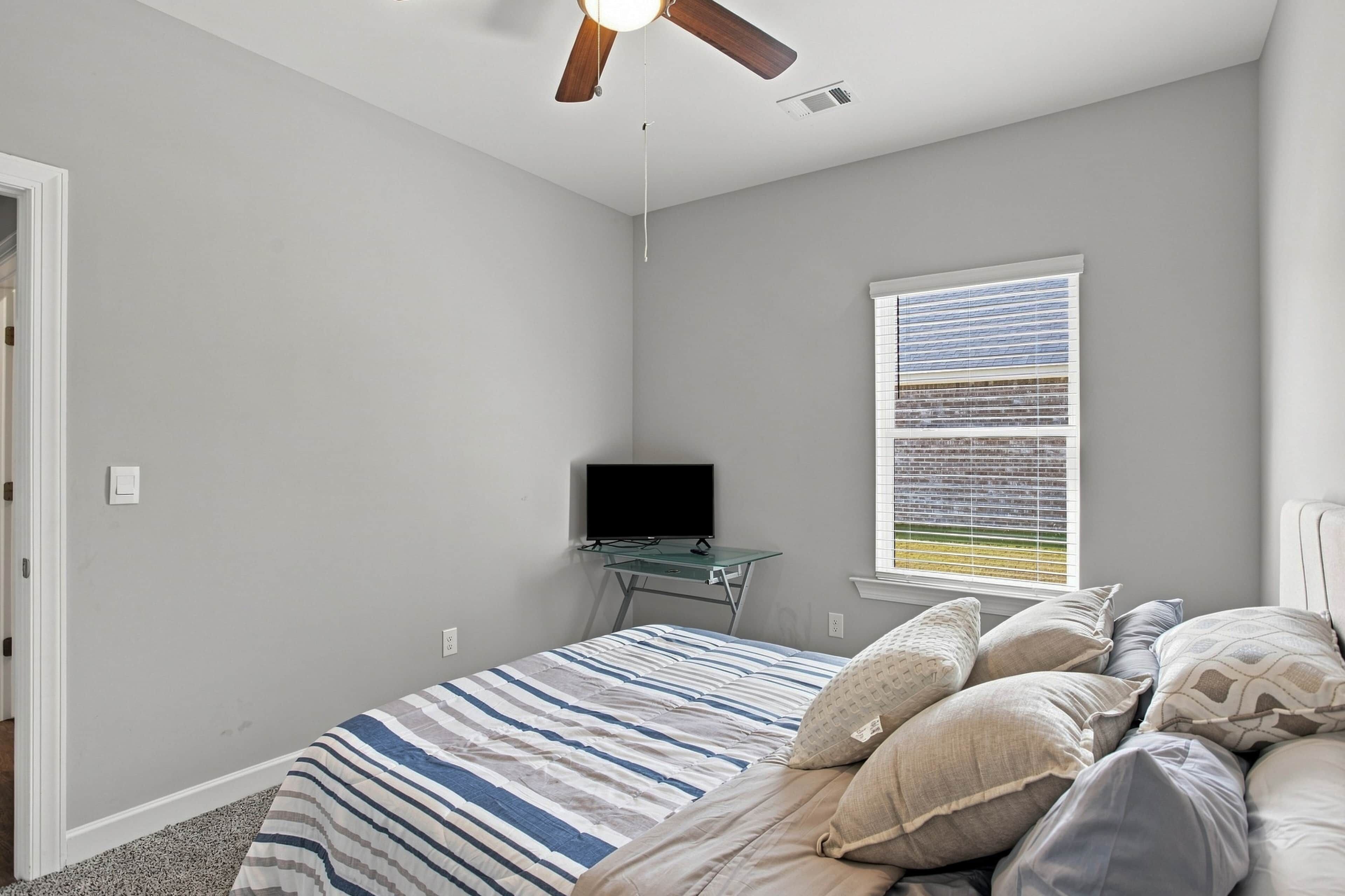 Un dormitorio con cama, televisor sobre un mueble, ventilador de techo, ventana con persianas y una puerta.