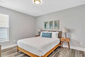 A bedroom with a bed, two bedside tables, a window with blinds, and three wall-mounted artworks.
