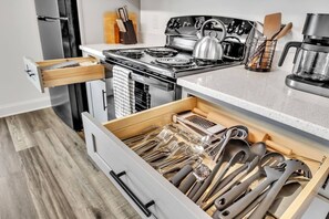Una cocina con estufa, hervidor y un cajón con diferentes utensilios.