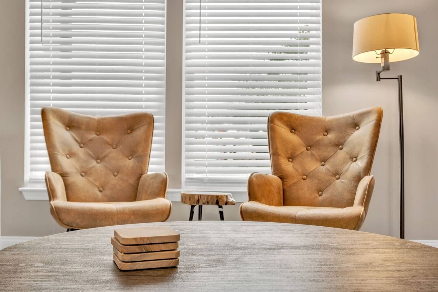 Two tufted armchairs facing each other with a round wooden table in between, set against a backdrop of white blinds and a floor lamp.