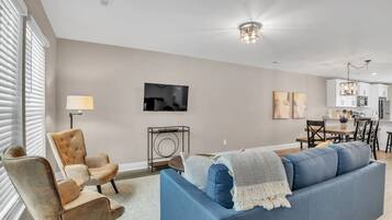 A modern living room with a blue sofa, a brown armchair, a TV, and a dining area with a table and chairs.