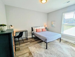 Un dormitorio con cama, mesita de noche, silla, escritorio y ventana con persianas.