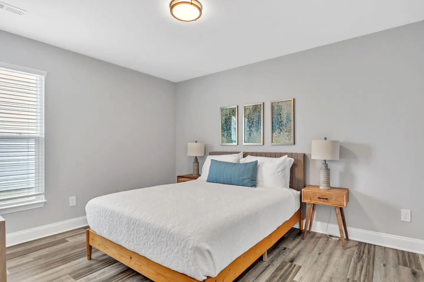A bedroom with a bed, two bedside tables, a window with blinds, and three wall-mounted artworks.