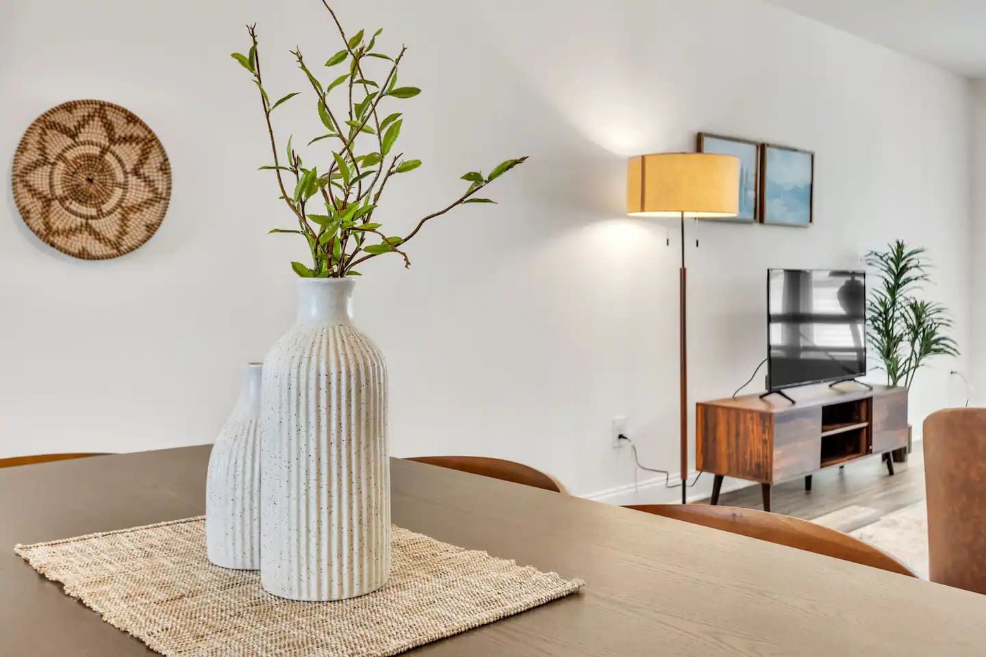 A modern living room with a dining table, a white vase with green branches, a floor lamp, a TV, and a wooden cabinet.