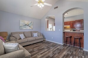 A living room with a sofa, a ceiling fan, and a kitchen area with wooden cabinets and a bar stool.