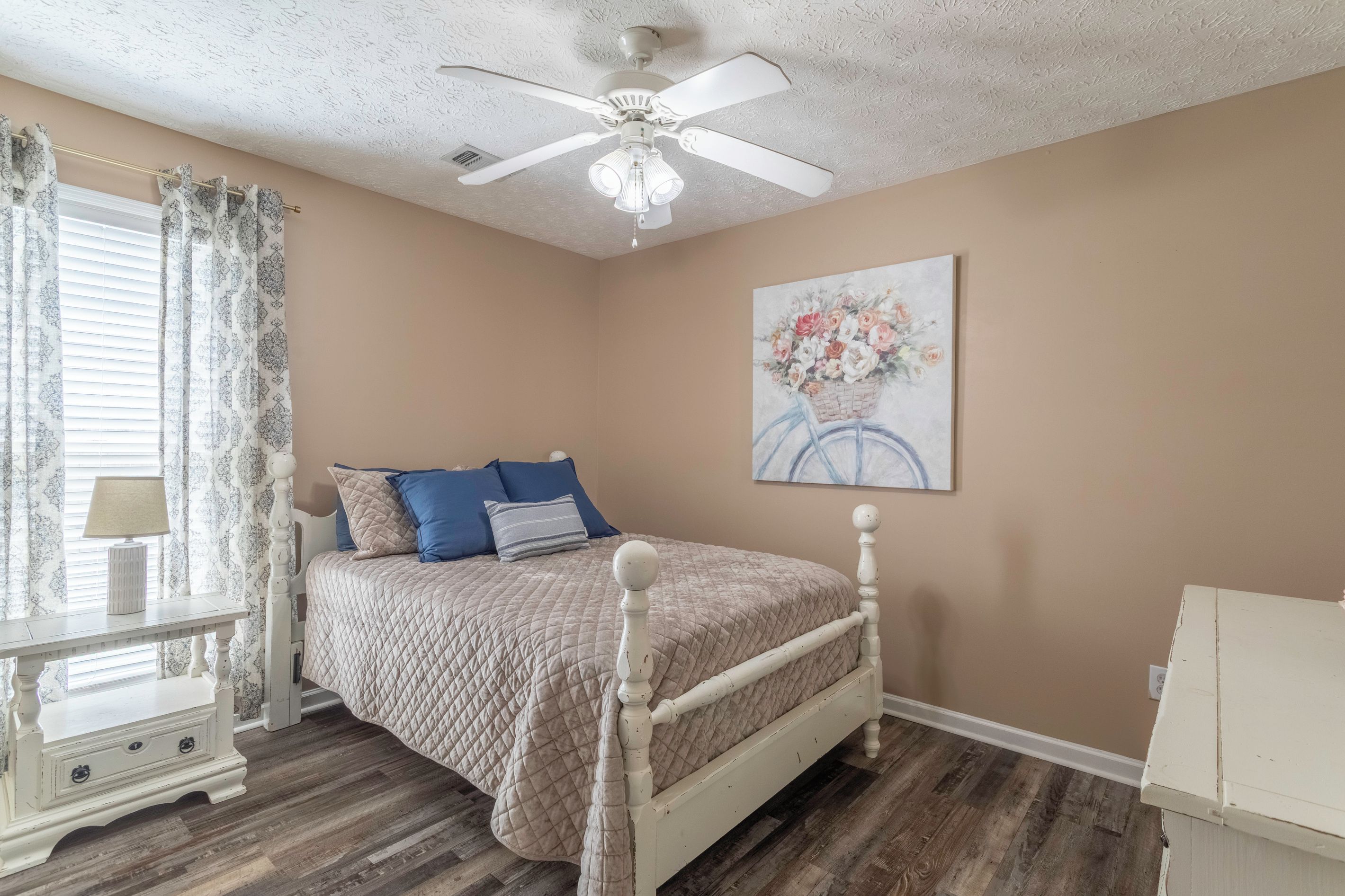 Une chambre à coucher avec un lit, une table de chevet, une lampe, un ventilateur de plafond et un tableau accroché au mur.