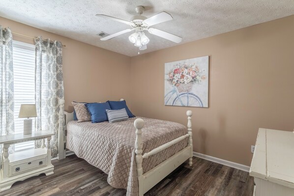A bedroom with a bed, a nightstand, a lamp, a ceiling fan, and a painting on the wall.