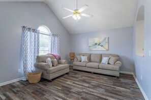 A living room with a ceiling fan, two sofas, a lamp, a painting, and a window with patterned curtains.