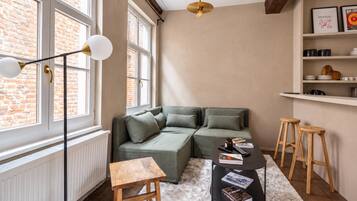 Un salon chaleureux avec un canapé d’angle, une petite table en bois et une table basse sur laquelle sont posés des livres et une télécommande.
