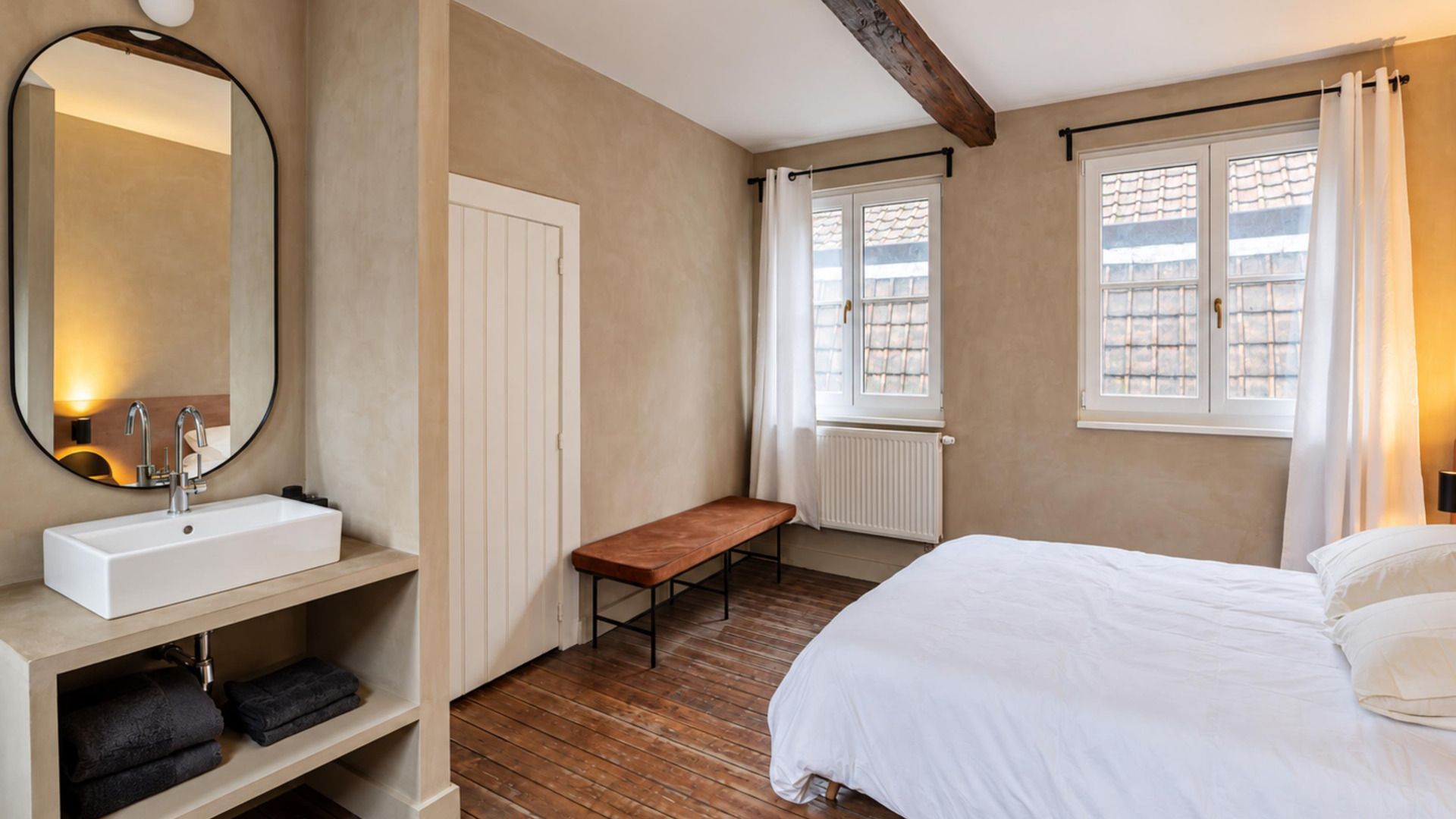 A bedroom with a bed, a bench, a bedside table with a mirror, and a window with curtains.