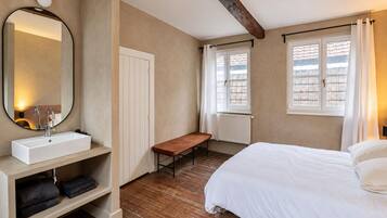 A bedroom with a bed, a bench, a bedside table with a mirror, and a window with curtains.