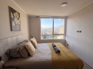 Een slaapkamer met een bed, kussens, een dekbed, een schilderij aan de muur en een raam met uitzicht op de stad.