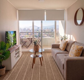 A living room with a sofa, a coffee table, a TV, and a view of a cityscape through large windows. - Edificio Byte Ñuñoa by Domus Travels (Ñuñoa)