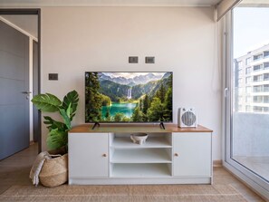 A flat-screen TV on a white entertainment unit with a mountainous landscape image, a white speaker, and a large green plant beside it. - Edificio Byte Ñuñoa by Domus Travels (Ñuñoa)