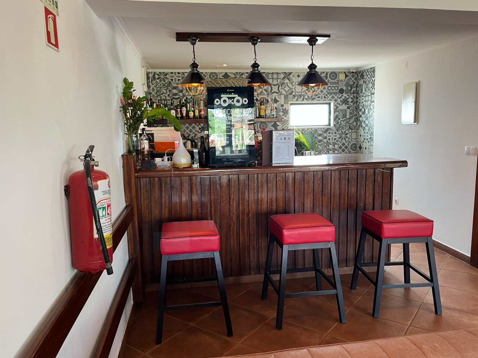 Una zona de bar con mostrador de madera, tres taburetes rojos, lámparas colgantes y un extintor en la pared.