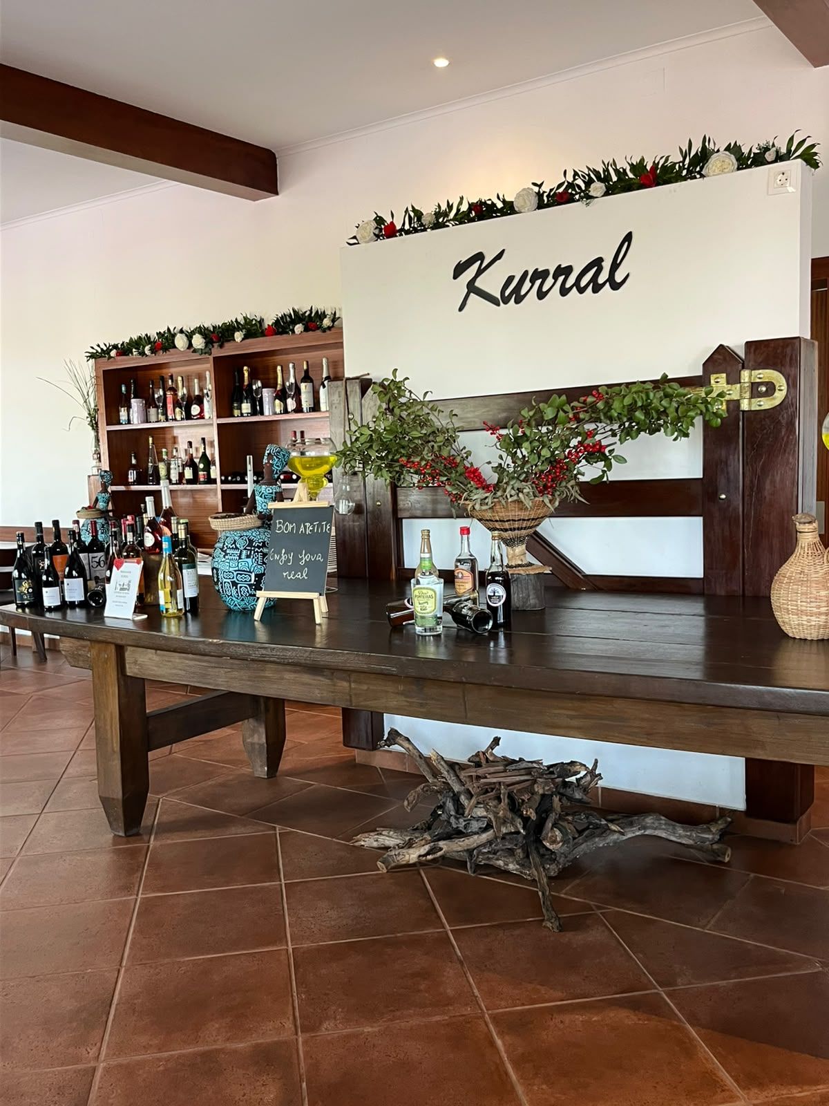 Un espace bar avec un comptoir en bois, des étagères remplies de bouteilles et une enseigne sur laquelle on peut lire « Kurral ».