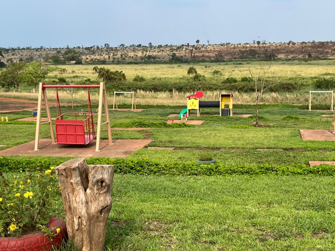 A playground with swings and a slide, surrounded by grassy fields and a hill in the background.