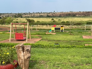 A playground with swings and a slide, surrounded by grassy fields and a hill in the background.