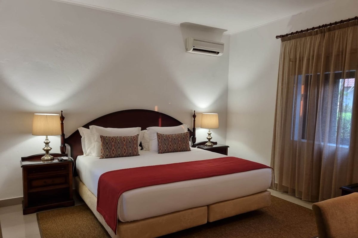 A hotel room with a large bed, two bedside tables with lamps, an air conditioning unit, and a window with curtains.