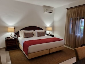 A hotel room with a large bed, two bedside tables with lamps, an air conditioning unit, and a window with curtains.