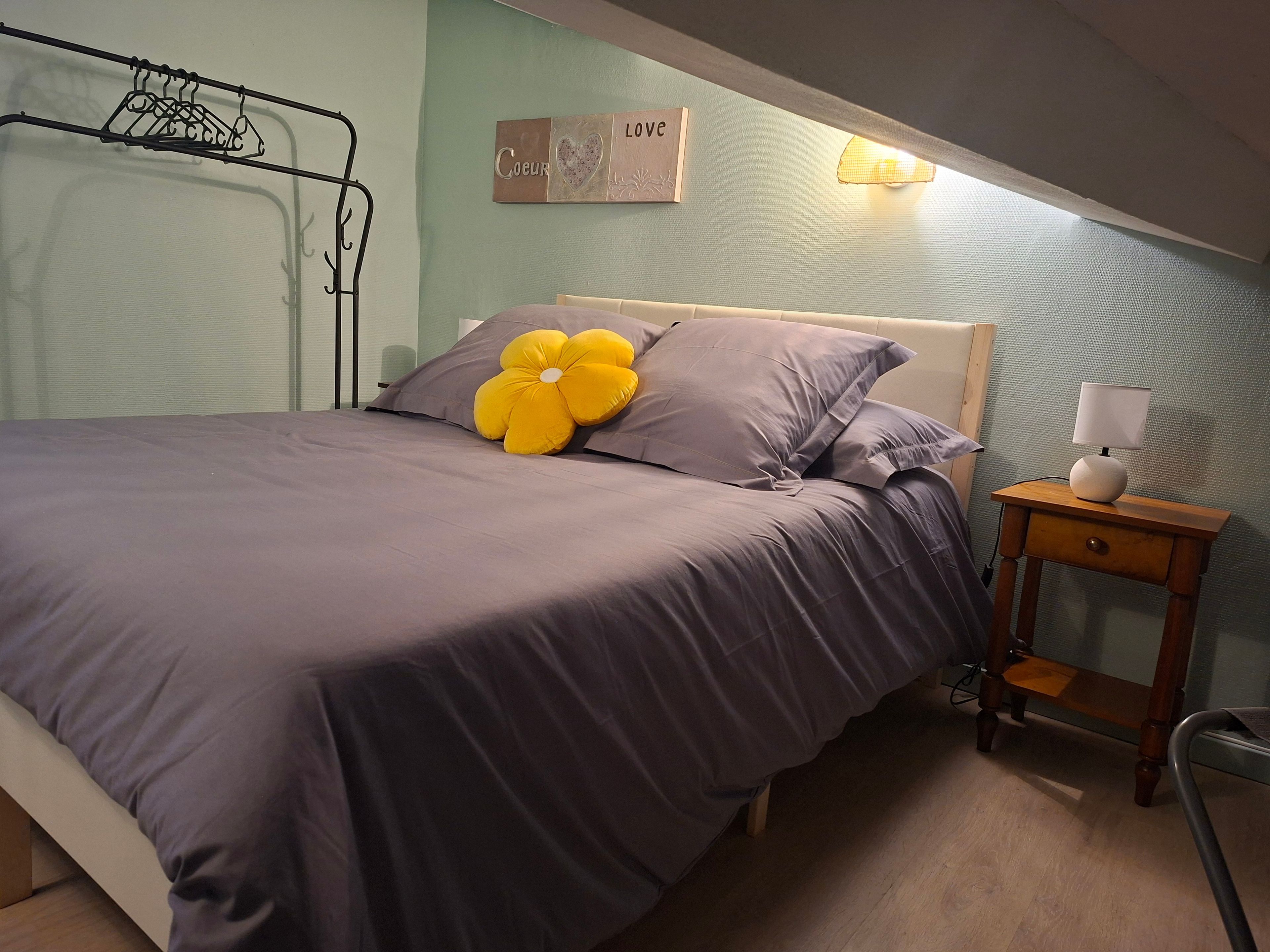 Un lit bien fait, agrémenté d’un grand coussin en forme de fleur, une table de chevet avec une lampe et un mur décoré d’un motif en forme de cœur.