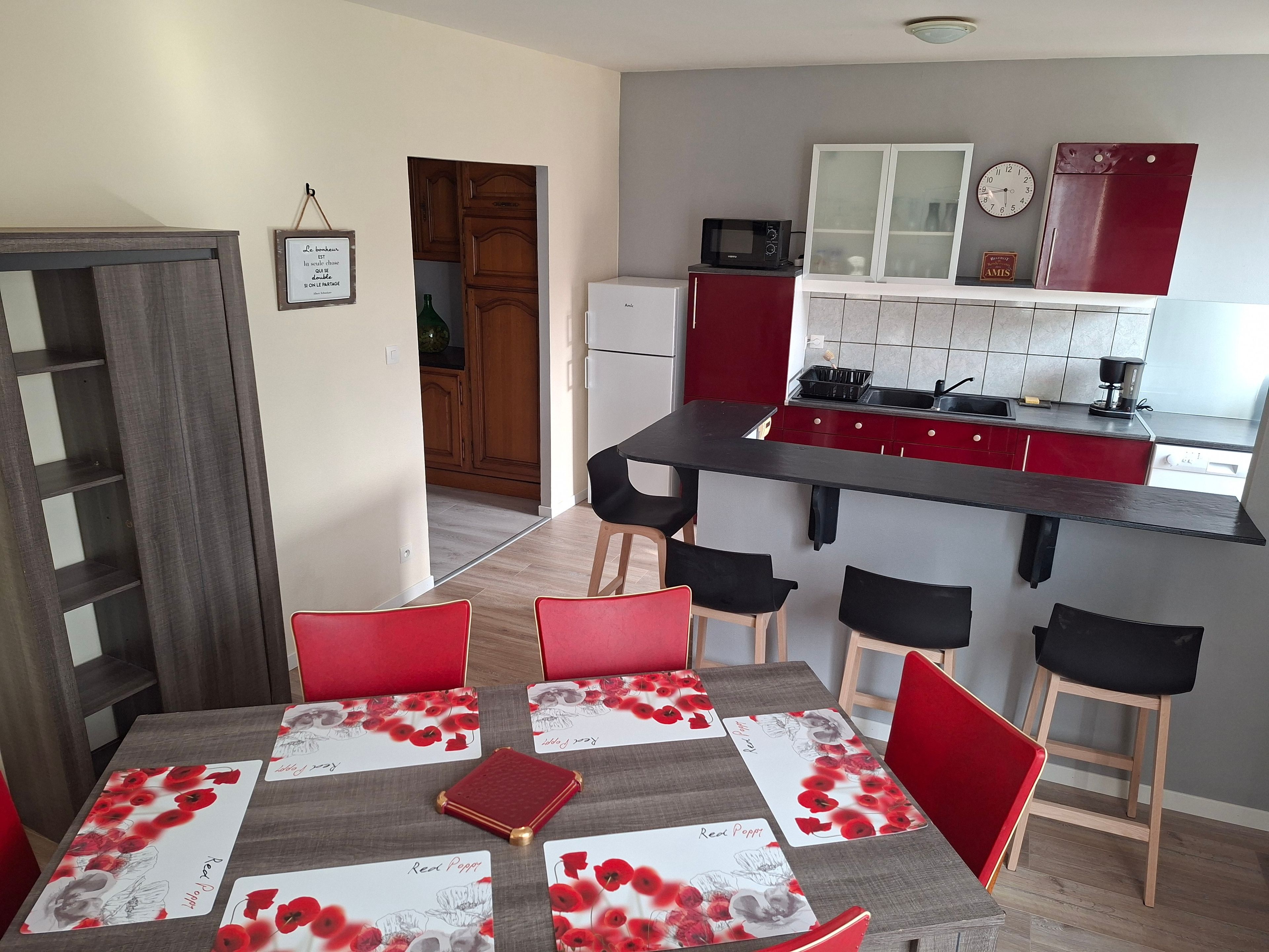 Un coin repas avec une table dressée pour quatre personnes, une cuisine avec des placards rouges, un four à micro-ondes et une horloge.