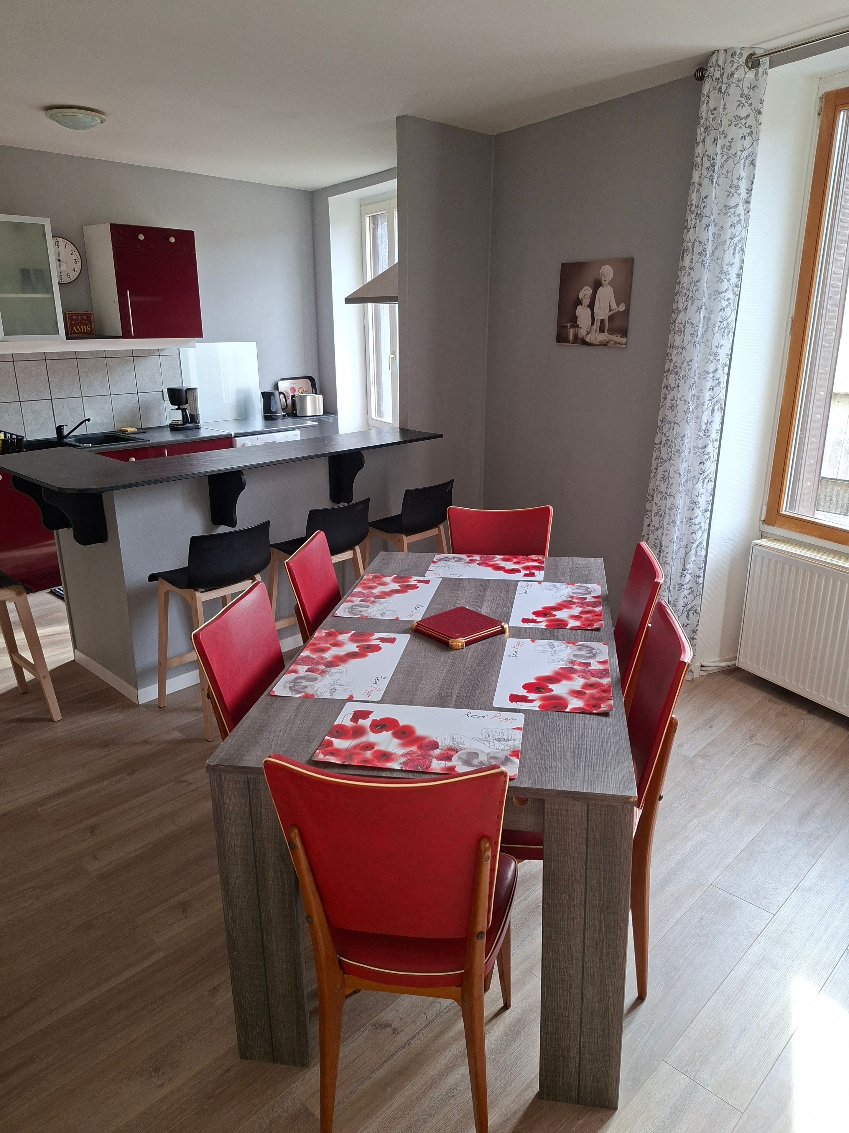 Ein Essbereich mit einem für vier Personen gedeckten Tisch, umgeben von roten Stühlen, eine Küche im Hintergrund und ein Fenster mit Vorhängen.
