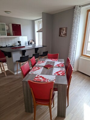 A dining area with a table set for four, surrounded by red chairs, a kitchen in the background, and a window with curtains.