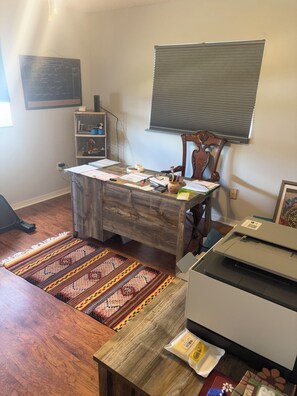 A room with a wooden desk, a printer, a chair, a window with blinds, a shelf with decorative items, and a patterned rug. - Adjacent to The Villages Property (Summerfield)